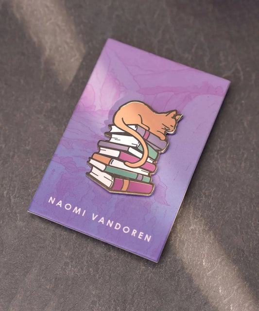 Enamel pin featuring a cute cat lounging on a stack of books by Naomi VanDoren, perfect for book lovers and cat enthusiasts.