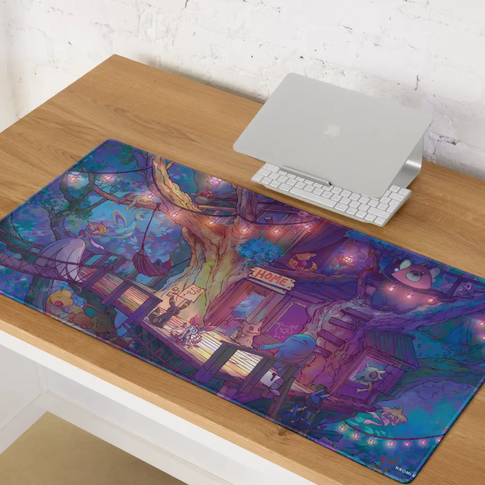 A large desk mat with a colorful fantasy forest illustration is laid out on a wooden desk next to a closed silver laptop and white keyboard. The illustration features purple, blue, and pink tones with whimsical trees, glowing lights, and small character figures.